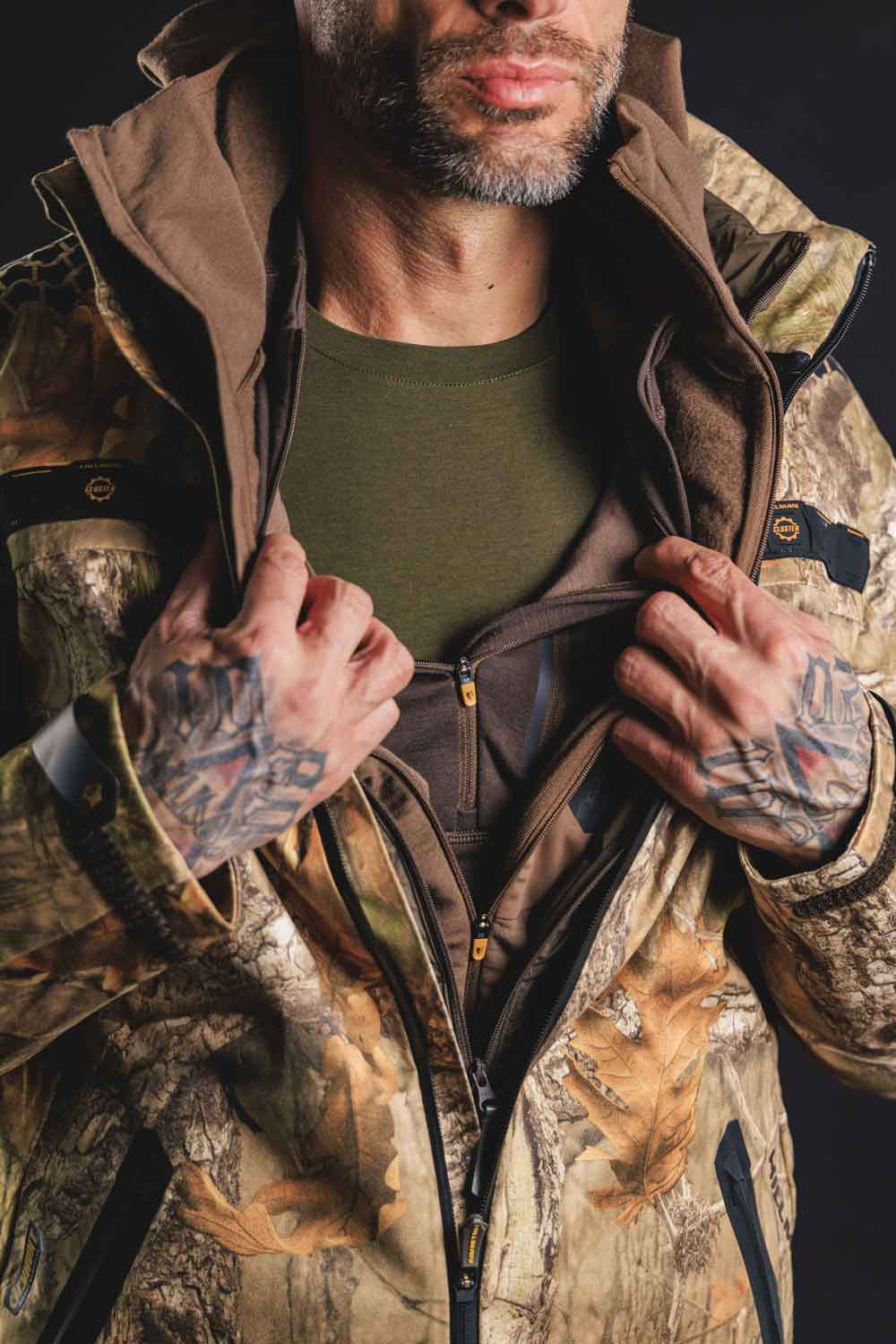 A person wearing a camouflage jacket with a brown hoodie and green shirt underneath. The individual is shown from the shoulders to the mouth, pulling the jacket open with tattooed hands. The background is dark.