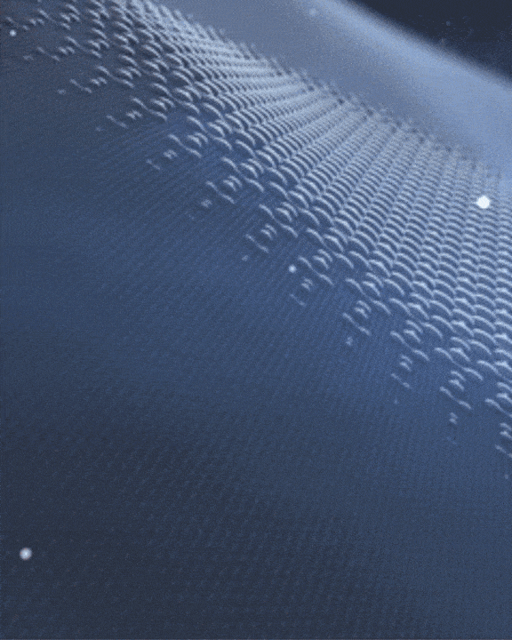 Close-up of a shiny, textured surface with a wavy pattern. Rows of small, raised bumps reflect light, creating a ripple effect across the material. The surface has a bluish tint and a smooth appearance.
