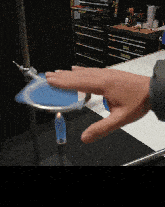 A person uses a blue paddle to levitate and move a small object. The setup is on a black surface with shelves and items in the background.