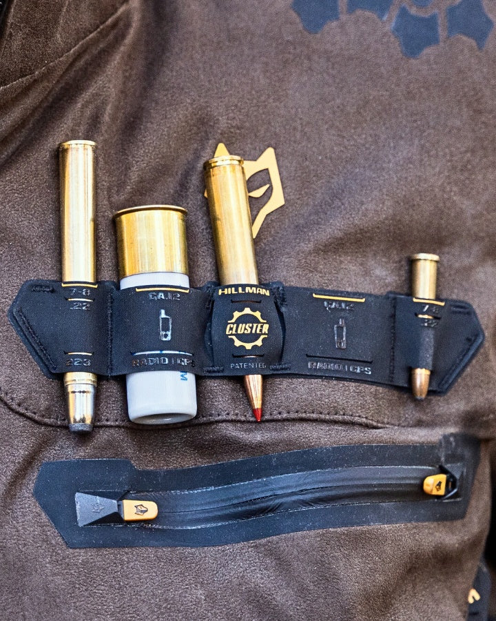 A leather belt with a holder on it containing three metallic cylindrical objects resembling bullet cartridges. The belt is on a brown leather surface, with a zippered pocket below featuring a small metallic logo.