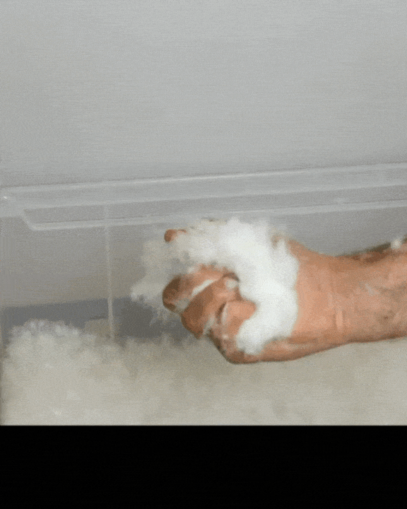 A hand compresses and releases cotton-like fluff near a transparent plastic container, demonstrating the materials softness and pliability.