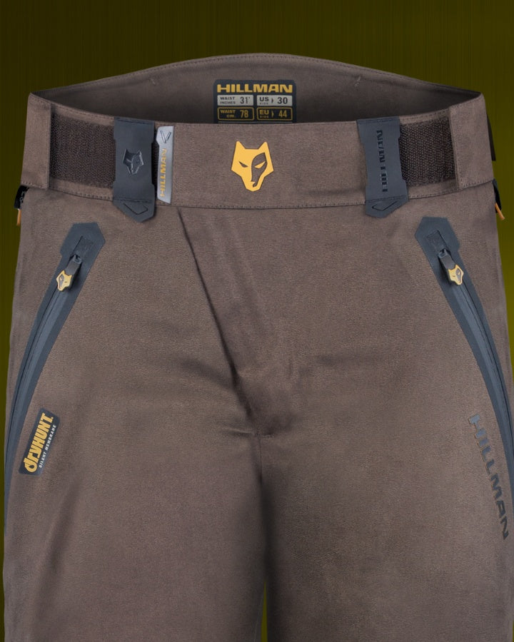 Close-up of brown outdoor pants with zipper pockets and belt loops. Theres a fox logo on the waistband and the brand name Hillman is visible on the pants.