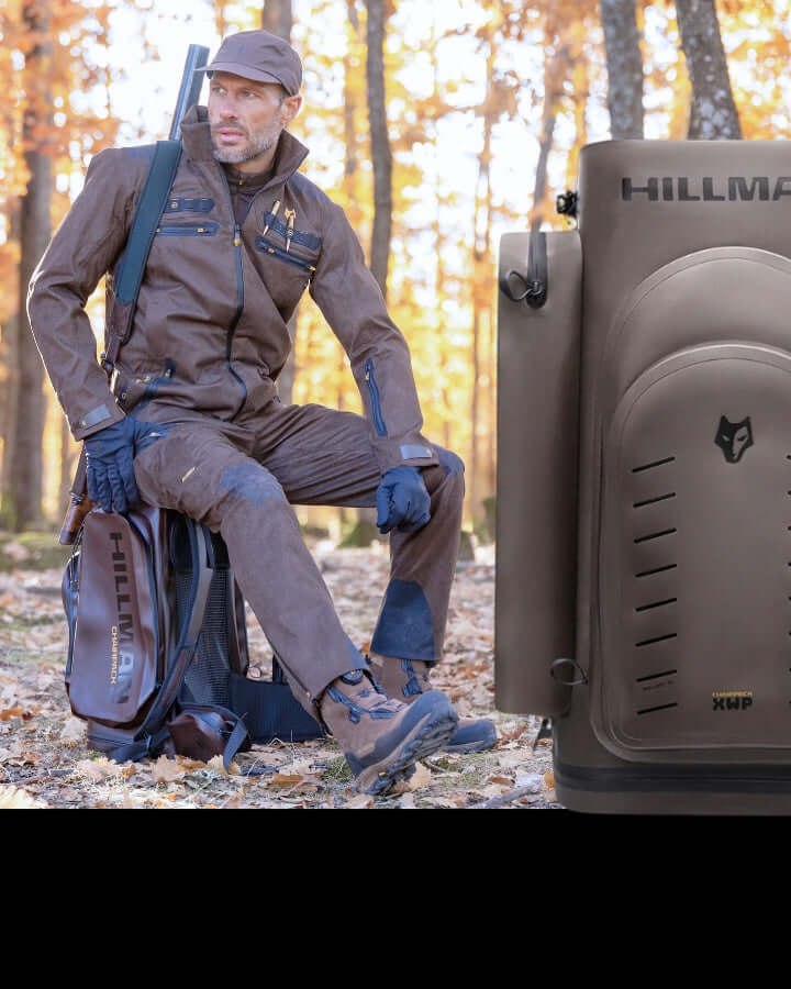 A person in brown outdoor gear stands in a forest during autumn. They have a backpack and a hat, and hold a water bottle. Nearby is a close-up of a HILLMAN backpack with a logo and zippers. Leaves cover the ground.