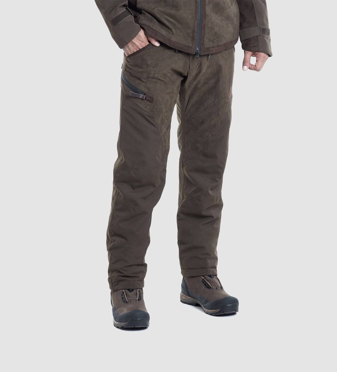 A person stands against a plain light gray background wearing a brown jacket, dark brown hiking boots, and Hillman® Waterproof Insulated Hunting Pants Fusion with a zippered pocket on the left thigh.