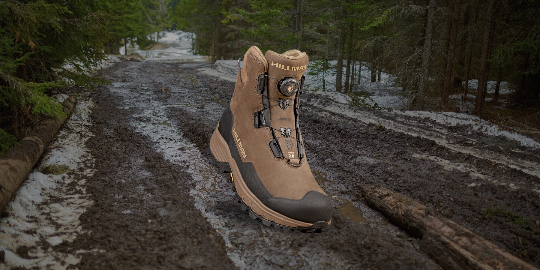 A single brown hiking boot is placed on a muddy forest trail surrounded by trees with patches of snow on the ground. The boot is upright, displaying its laces and rugged sole.