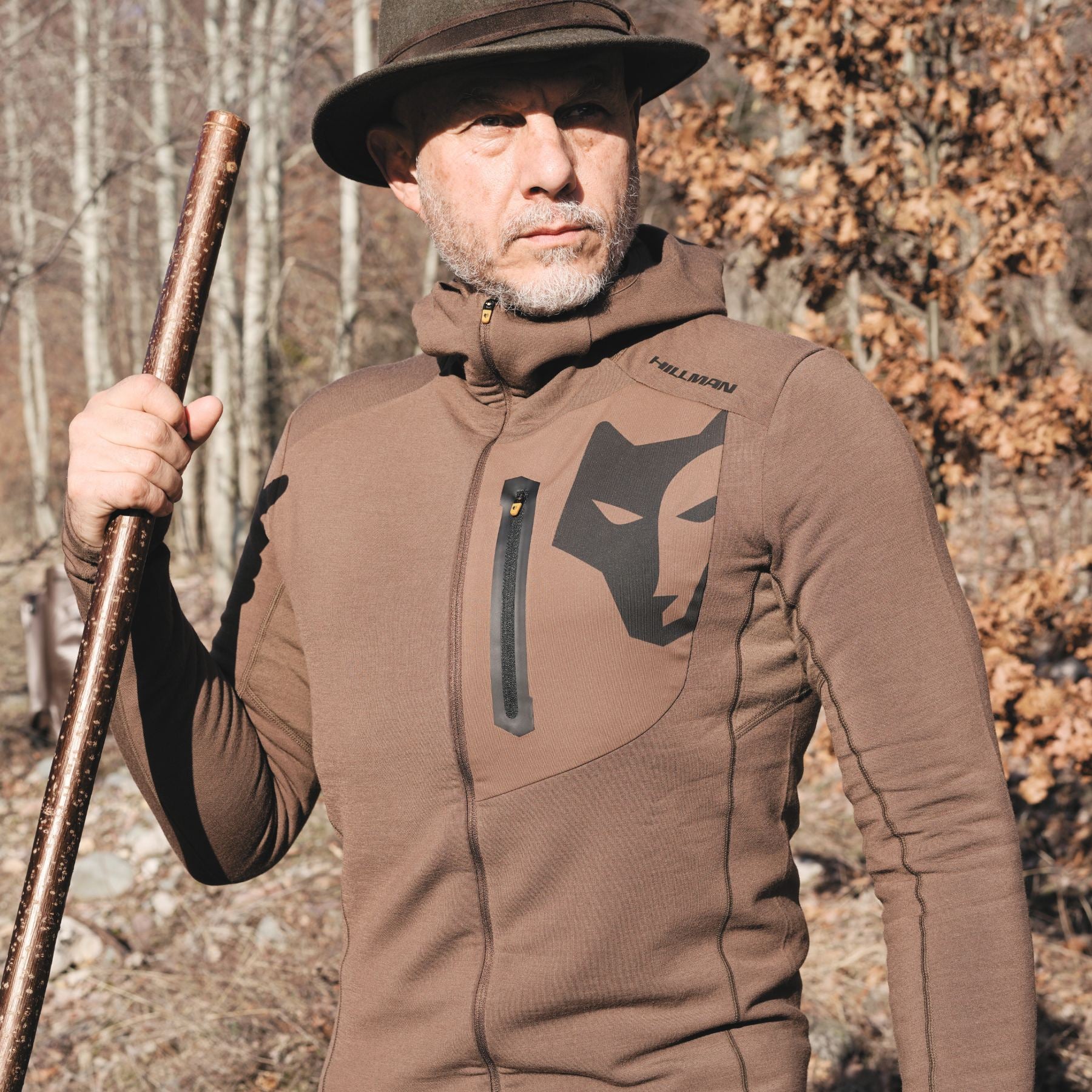 A man wearing the Hillman® Hunting Hoodie | Merino Wool & Bamboo Fleece Mid Layer | 3ML, featuring a large wolf face graphic and zippered chest pocket, stands outdoors among dry trees holding a wooden staff.