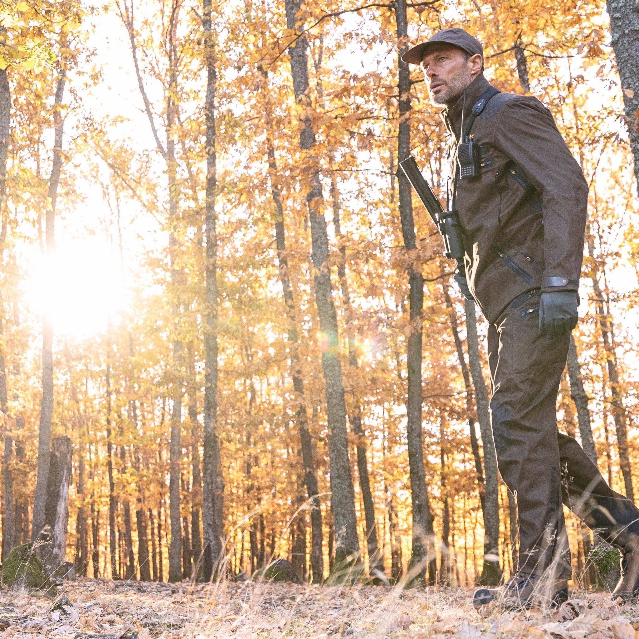 Wearing HILLMAN GEAR Insulated Hunting Pants 6OL Cold Weather Waterproof Bibs, a hunter with a cap and gloves walks through a sunlit autumn forest, rifle slung over their shoulder amid tall trees and fallen leaves.