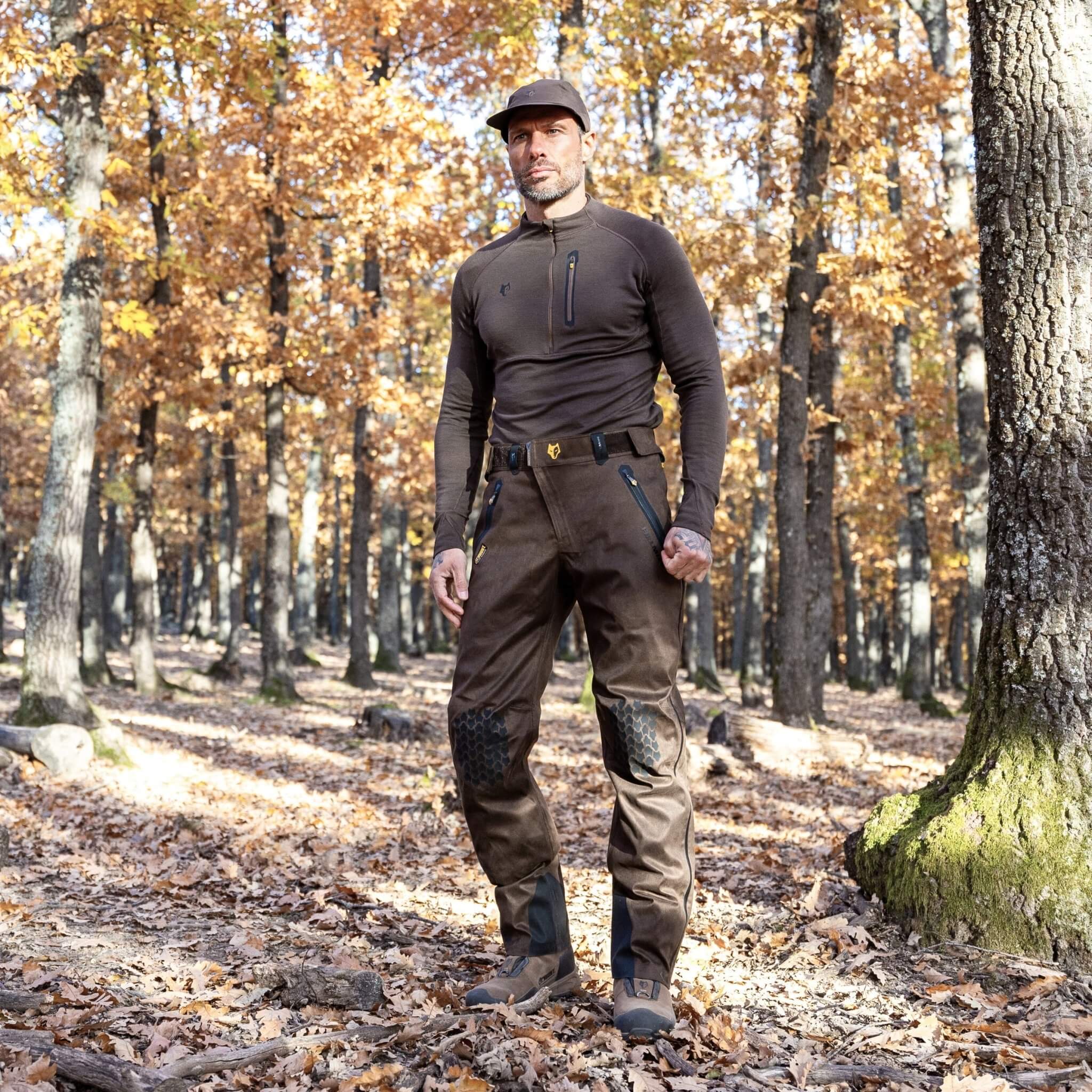 A man wearing Hillman® Waterproof Hunting Pants | Lightweight AlphaShell 5WL and a cap stands in a sunlit forest with autumn leaves on the ground and orange foliage in the background.