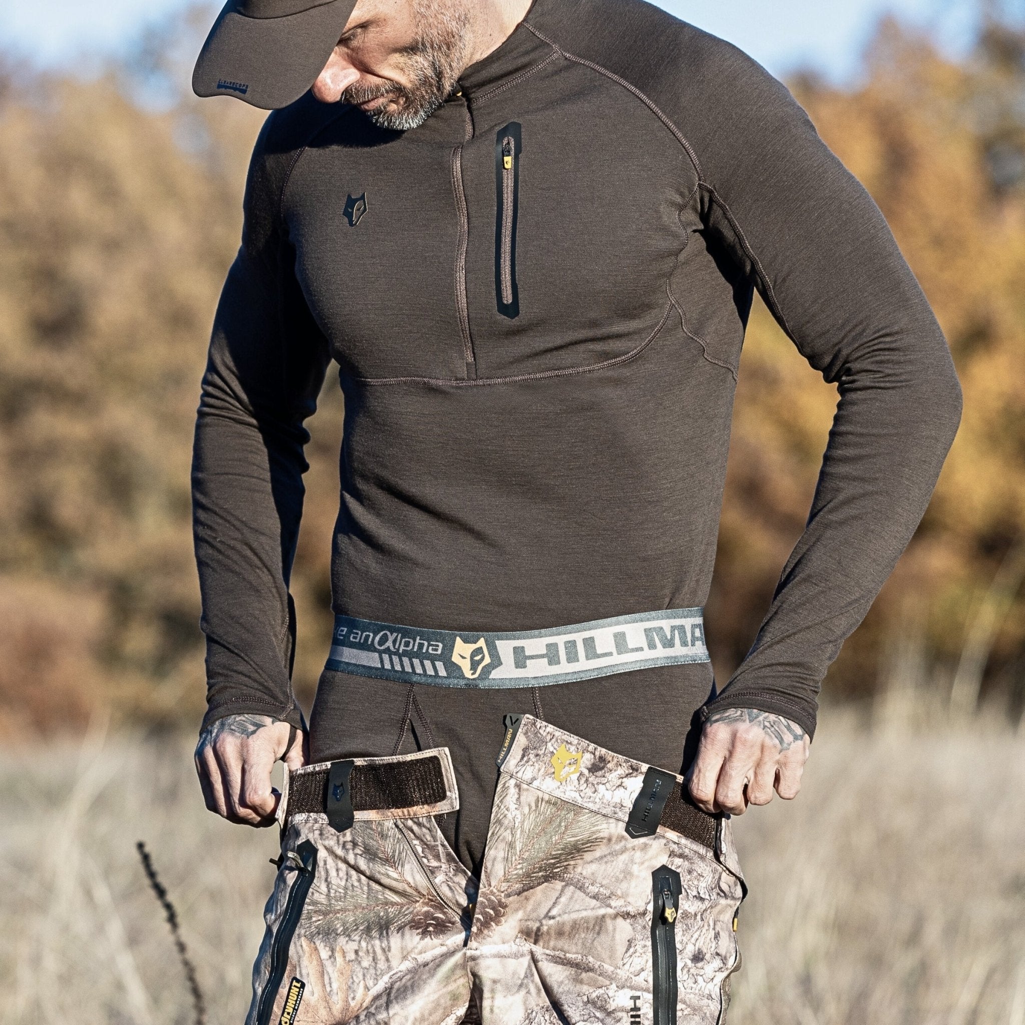 A man outdoors adjusts his waistband while wearing Hillman® Hunting Long Johns | Merino Wool Base Layer Bottom | 2BL with suspenders. Dry grass and blurred autumn trees appear in the background.