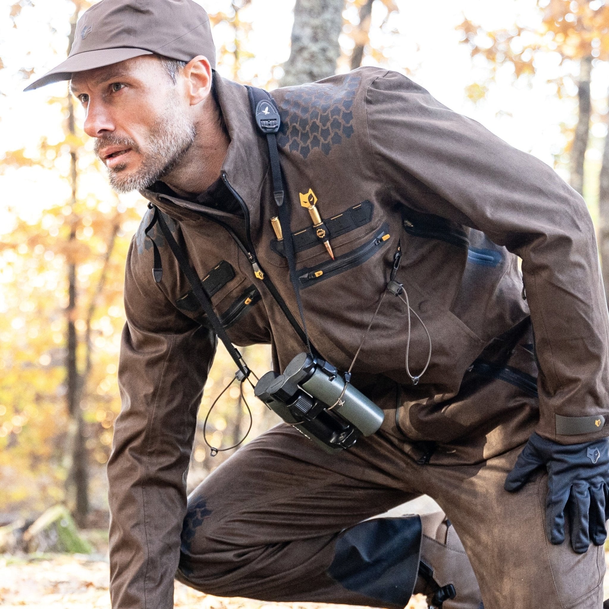 A man in a brown outfit and cap crouches in a forest, wearing gloves and the HILLMAN GEAR 6OL Cold Weather Camo Hunting Jacket - Heated Camouflage Coat. Binoculars hang around his neck, blending perfectly into the autumn foliage, ready for adventure.