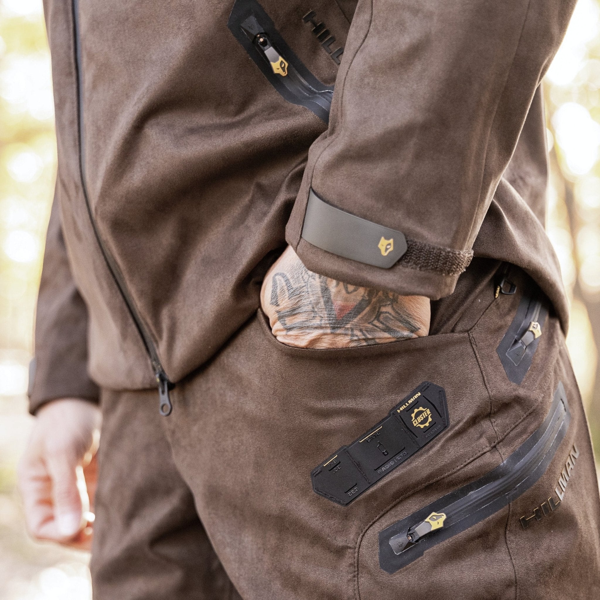 A person wearing HILLMAN GEAR Insulated Hunting Pants 6OL Cold Weather Waterproof Bibs and a matching jacket with black and yellow accents stands outdoors, one tattooed hand partially visible in their pocket.