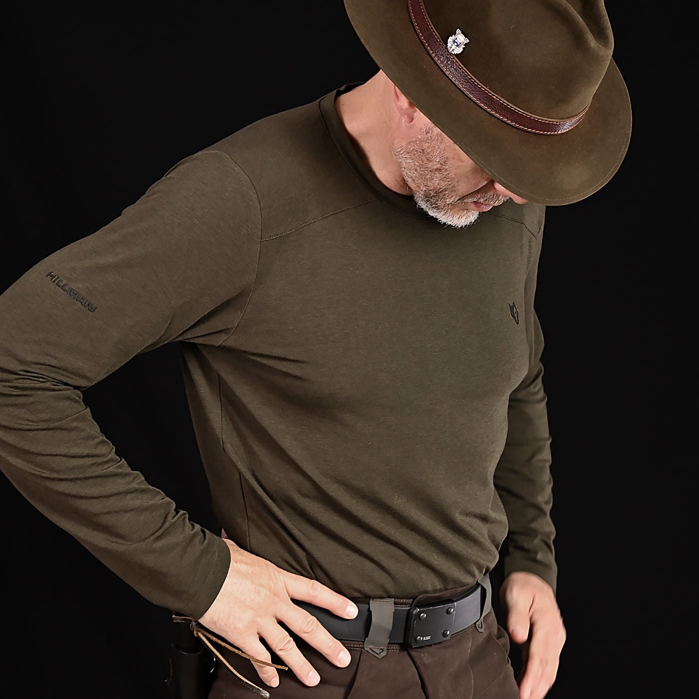 A man wearing a brown hat, brown pants, and a Hillman® 1UL Bamboo Long Sleeve Hunting T-Shirt—an ultralight all-season base layer—stands against a black background, looking down with one hand on his hip.