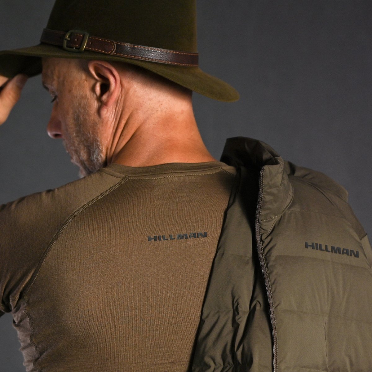 A man in a brown shirt and green hat adjusts his hat. He has a Hillman® 4IL Hunting Down Jacket - Packable Puffy Coat - 1000FP Puffer draped over his shoulder, matching his shirt.