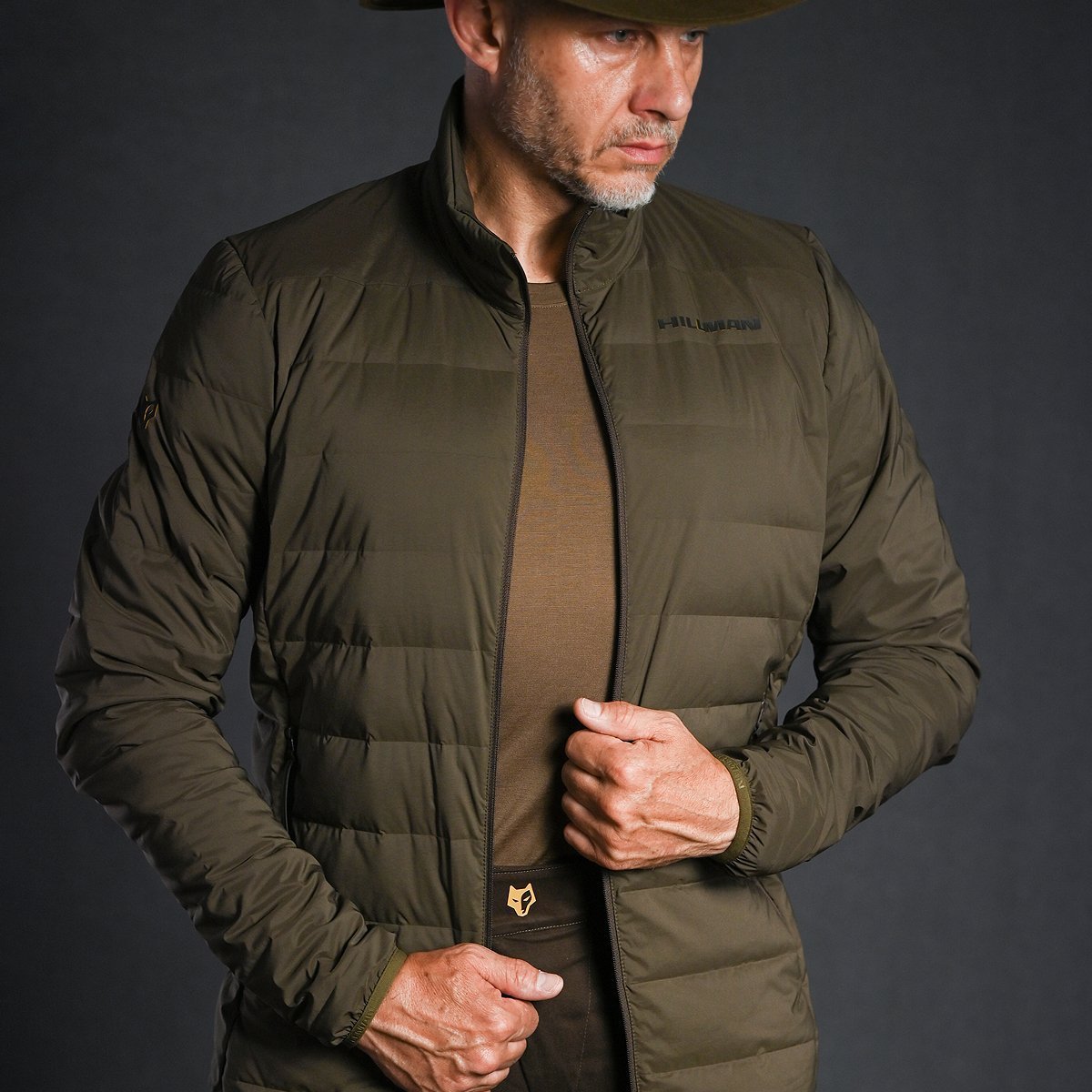 A man in a Hillman® 4IL Hunting Down Jacket – Packable Puffy Coat (1000FP Puffer), worn over a brown shirt, stands against a dark background, holding his jacket and looking to the side while wearing a wide-brimmed hat.