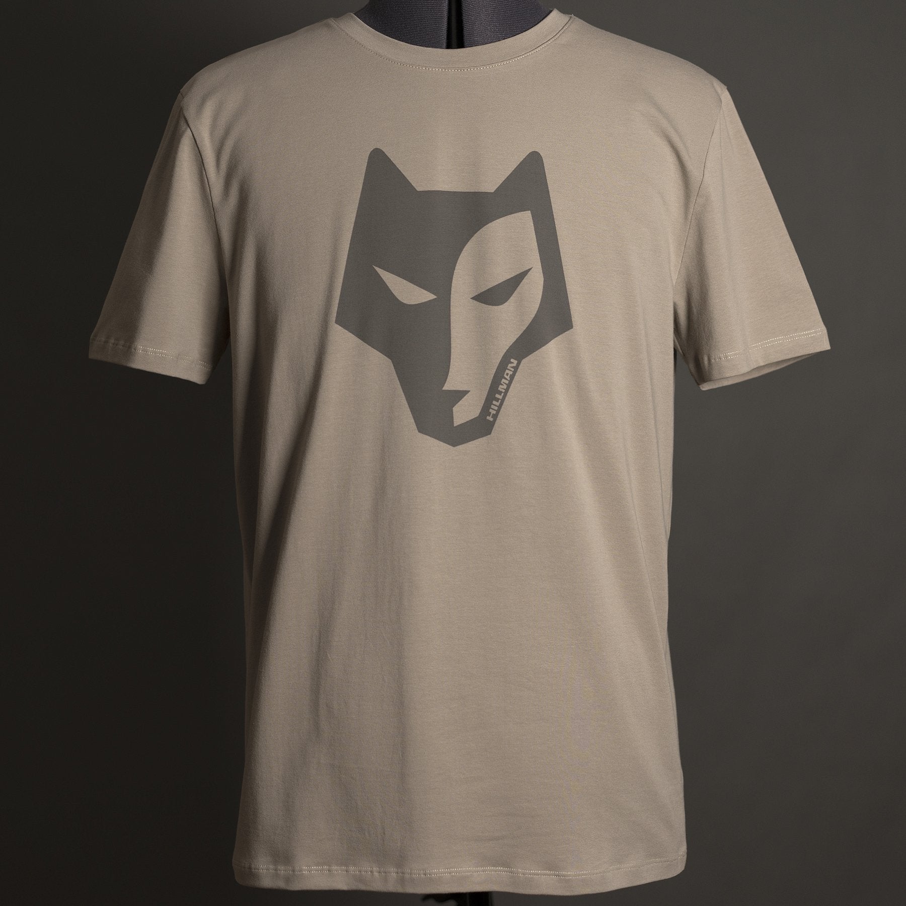 Introducing the Alpha T-shirt by Hillman - България: a plain beige tee crafted from luxurious Egyptian cotton, featuring a stylized dark gray wolf head logo on the front. Displayed on a mannequin against a dark background, this shirt embodies both style and comfort.