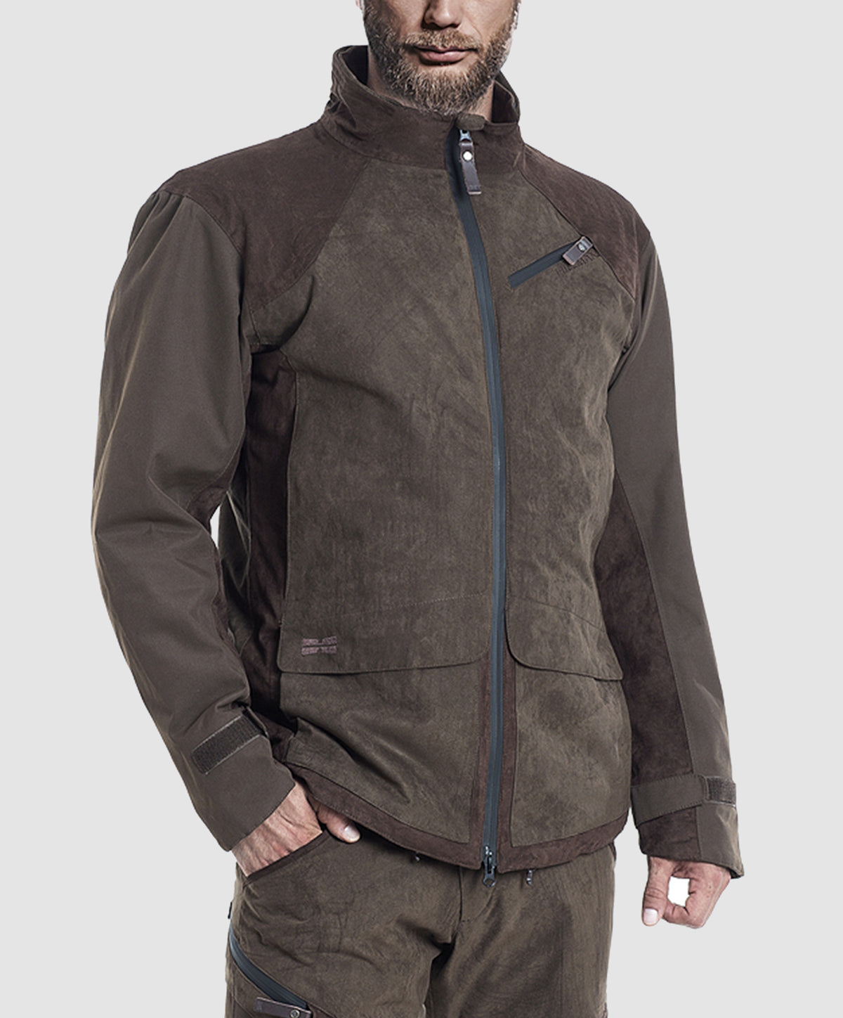 A man wearing the Hillman® PRE-ORDER Fusion Hunting Jacket, a durable brown outdoor piece with a high collar, zippered front, and chest pocket, along with matching pants, stands against a plain light gray background.