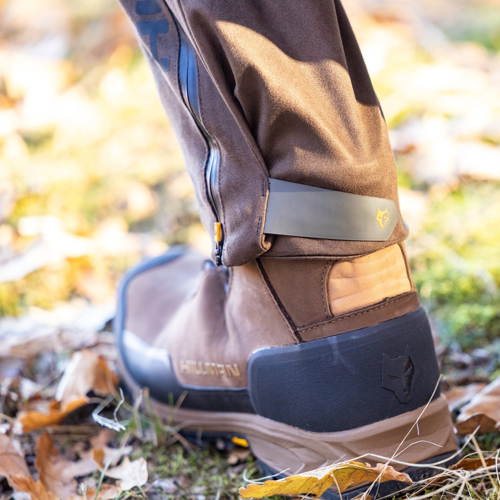 Close-up of a person wearing HILLMAN GEAR Insulated Hunting Pants 6OL Cold Weather Waterproof Bibs tucked into brown outdoor boots with wolf head logos, standing on grass and fallen leaves.