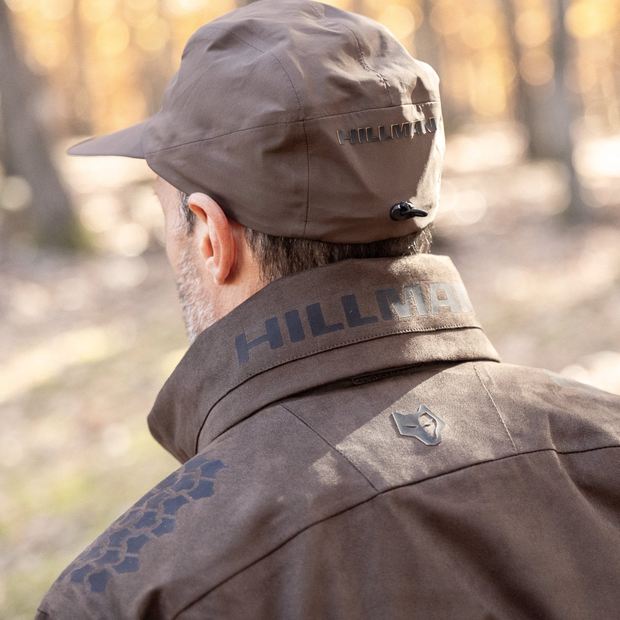 A person wears the HILLMAN GEAR Waterproof Hunting Jacket 6OL Heated-Extreme Cold Insulated and matching cap, both branded “HILLMAN,” standing outdoors in a woodland with blurred trees, ready for extreme cold.