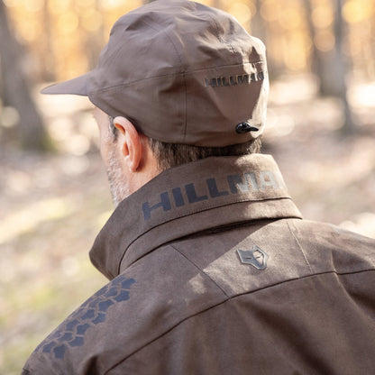 A person wears the HILLMAN GEAR Waterproof Hunting Jacket 6OL Heated-Extreme Cold Insulated and matching cap, both branded “HILLMAN,” standing outdoors in a woodland with blurred trees, ready for extreme cold.