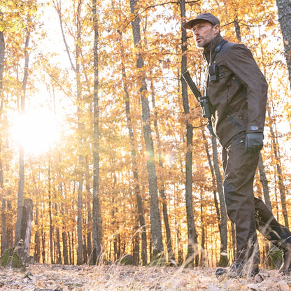 Wearing HILLMAN GEAR Insulated Hunting Pants 6OL Cold Weather Waterproof Bibs, a hunter with a cap and gloves walks through a sunlit autumn forest, rifle slung over their shoulder amid tall trees and fallen leaves.