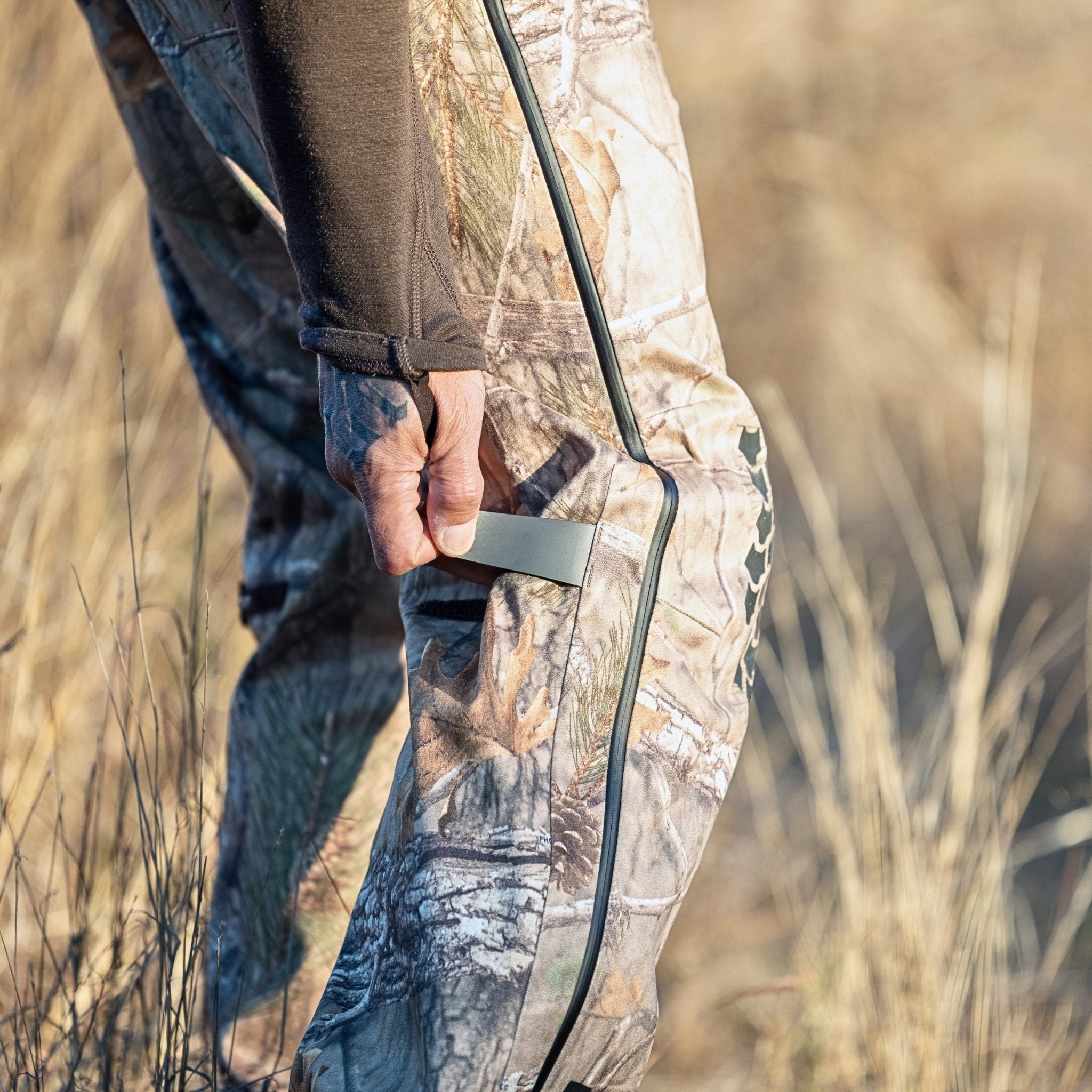Dressed in HILLMAN GEARs 5WL Ultra-Lightweight Waterproof Hunting Pants, they stand in the dry grass tugging at a zipper. Their long-sleeve shirt adds to the rural vibe, while the breathable fabric of their attire is crafted for practical outdoor adventures.