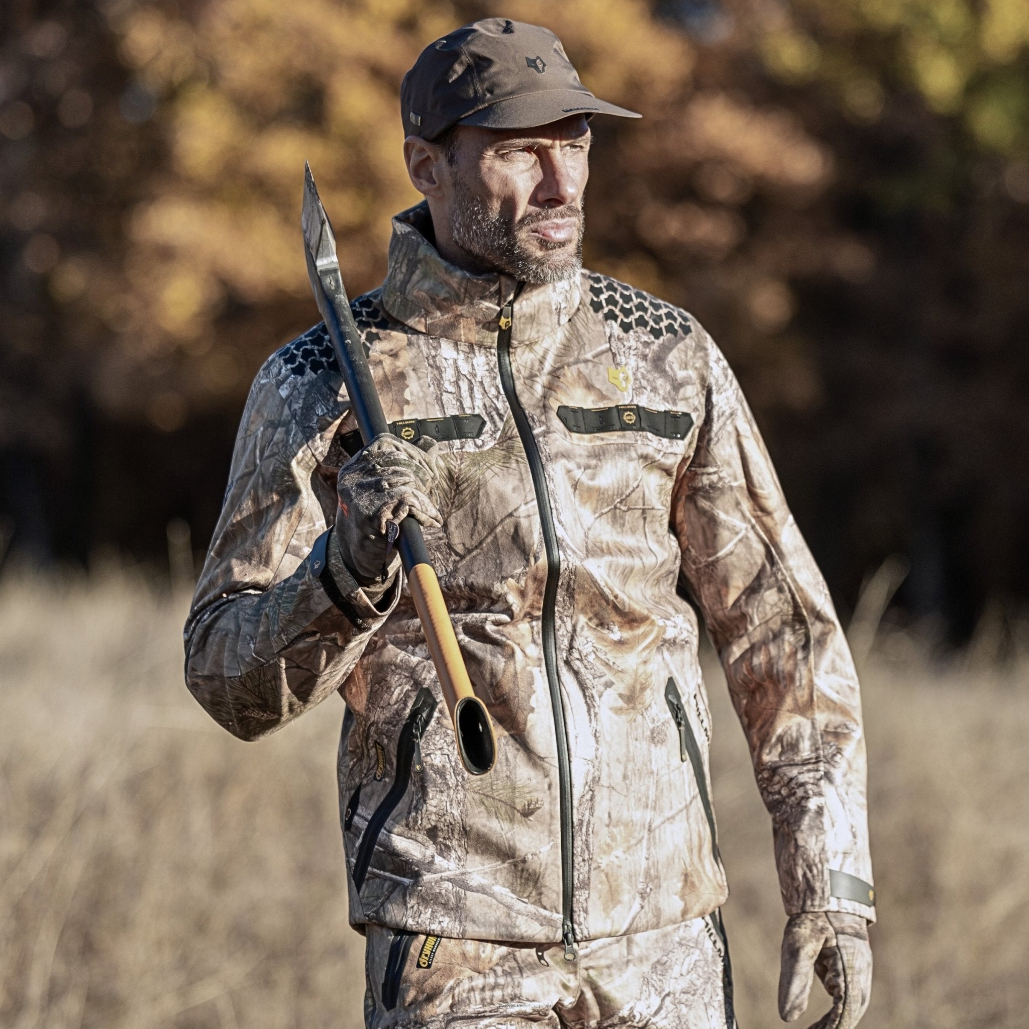 A person wearing the HILLMAN GEAR 5WL Waterproof Camo Hunting Rain Jacket stands outdoors with a pointed tool, blending into the natural setting of blurred foliage and grass.