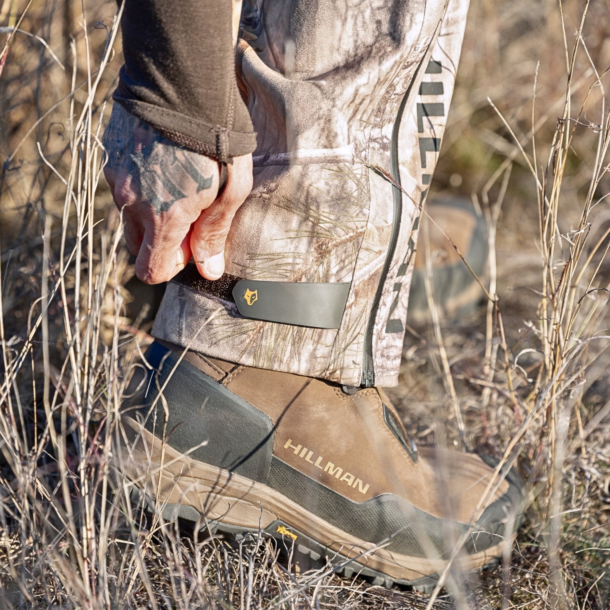 In a dry grassy area, someone wearing HILLMAN GEARs 5WL Ultra-Lightweight Waterproof Hunting Pants in camouflage adjusts a strap on their brown hiking boot. The pants are designed for comfort and mobility and perfect for any adventure, with the side view focusing on the lower leg and boot.