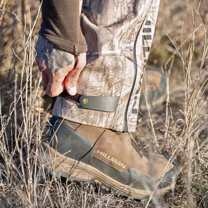 In a dry grassy area, someone wearing HILLMAN GEARs 5WL Ultra-Lightweight Waterproof Hunting Pants in camouflage adjusts a strap on their brown hiking boot. The pants are designed for comfort and mobility and perfect for any adventure, with the side view focusing on the lower leg and boot.