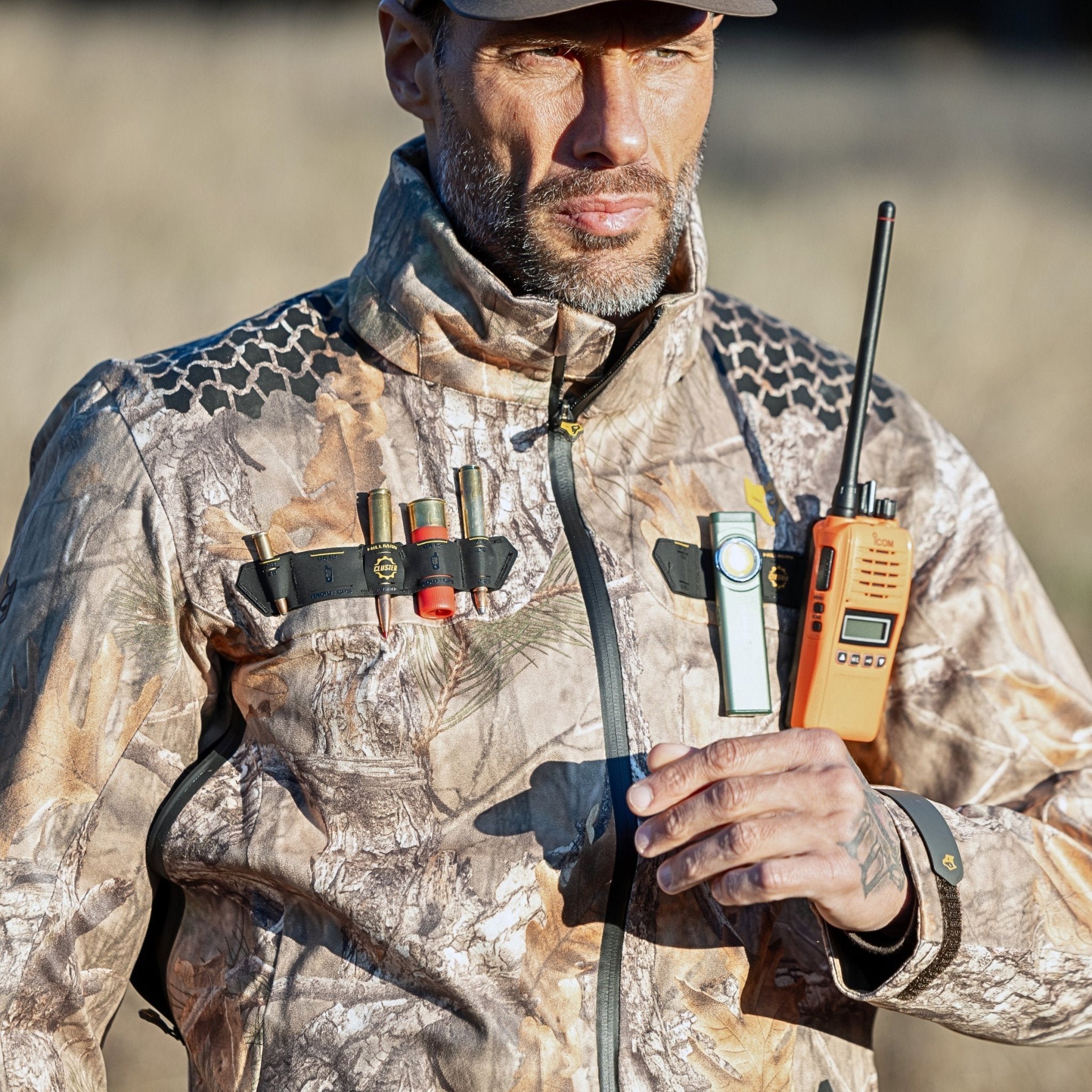 A man is outdoors wearing a HILLMAN GEAR 5WL Waterproof Camo Hunting Rain Jacket, with an orange two-way radio in his left hand and shotgun shells clipped to his jacket. He wears a cap, has a serious expression, and the background is blurred.