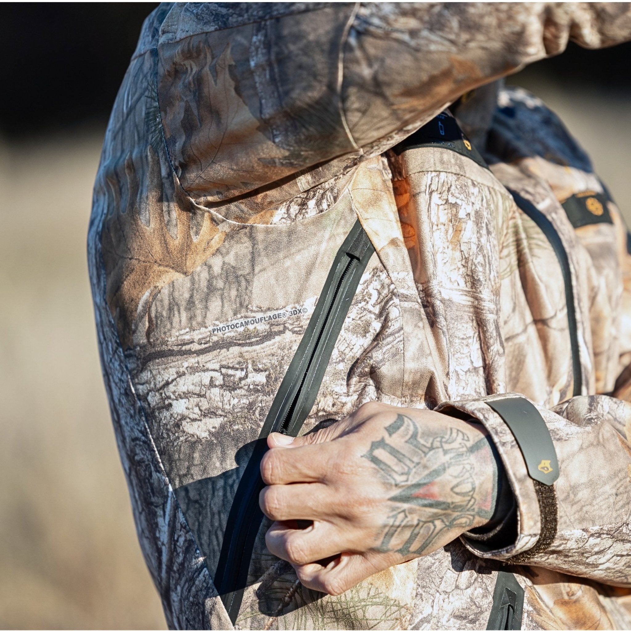 A person wearing the HILLMAN GEAR 5WL Waterproof Camo Hunting Rain Jacket zips a pocket. The ultra-light tree bark camo is perfect for outdoor activities, while tattoos are visible on the hand and a blurred field serves as the backdrop.