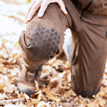 A person, shown from the waist down, kneels on fallen leaves wearing Hillman® Insulated Hunting Pants - Waterproof | AlphaShell 6OL, featuring brown fabric and a textured knee patch, with one hand resting on their knee.