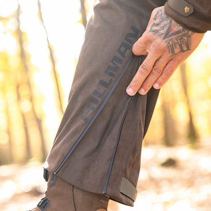 A close-up of a tattooed hand zipping up the HILLMAN GEAR Insulated Hunting Pants 6OL Cold Weather Waterproof Bibs, with HILLMAN branding and rugged brown footwear—ideal outdoor wear for staying warm and dry.