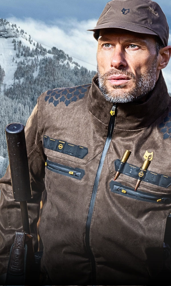 A man wearing a brown cap and jacket with black accents is holding a firearm. The snowy mountain landscape is visible in the background. The jacket has pockets with zippers and hooks. The man has a serious expression.