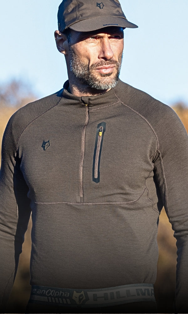 A man with a beard wearing a brown quarter-zip pullover and cap stands outdoors. The pullover features a small logo on the chest and a zippered pocket. The sky in the background is clear.