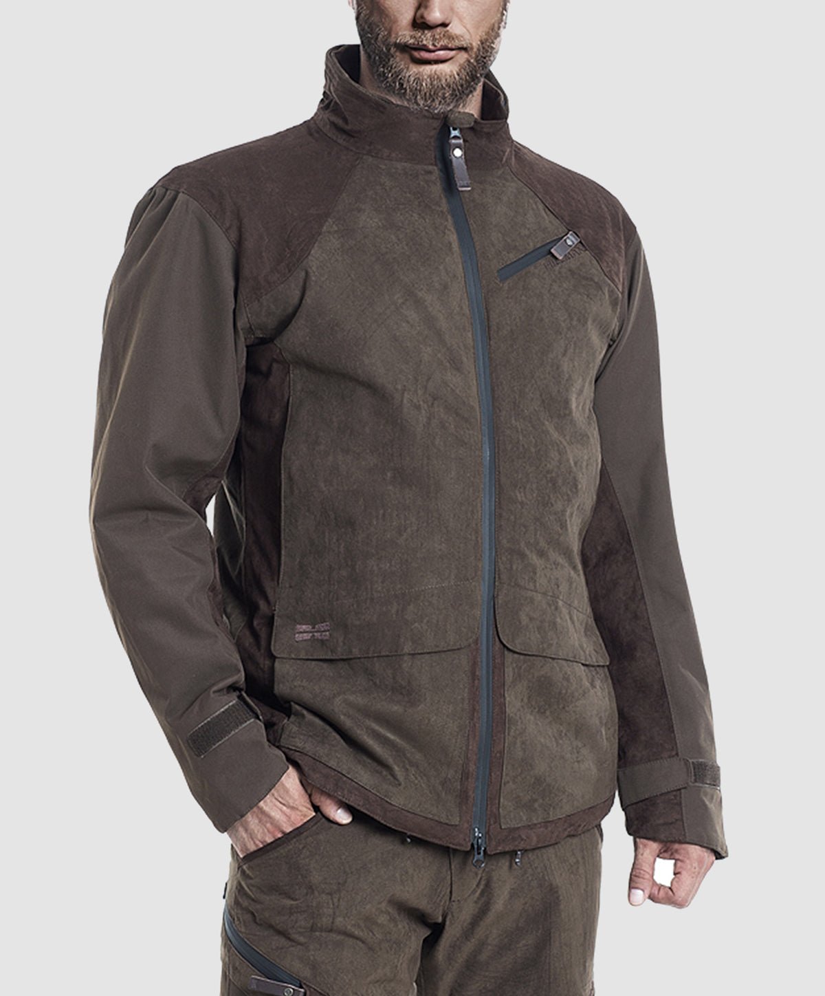 A man wearing the Hillman® PRE-ORDER Fusion Hunting Jacket, a durable brown outdoor piece with a high collar, zippered front, and chest pocket, along with matching pants, stands against a plain light gray background.