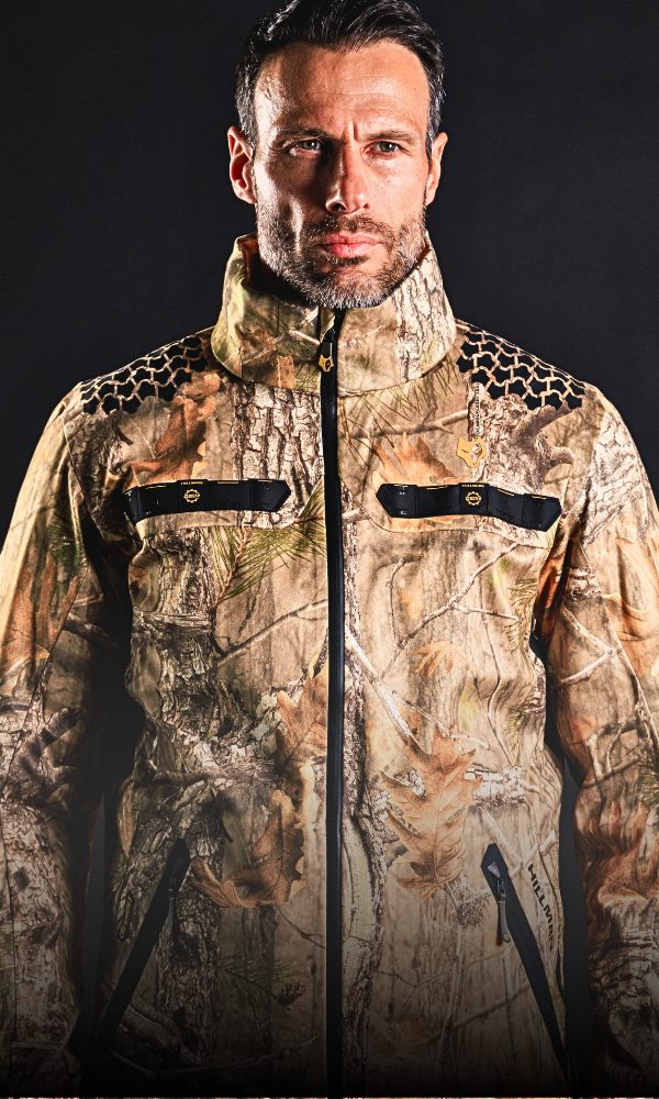 A man stands facing forward wearing a camouflage-patterned jacket with a high collar and zipped pockets. The jacket has a mix of earthy tones and black accents. The background is dark and plain, highlighting the jackets details.