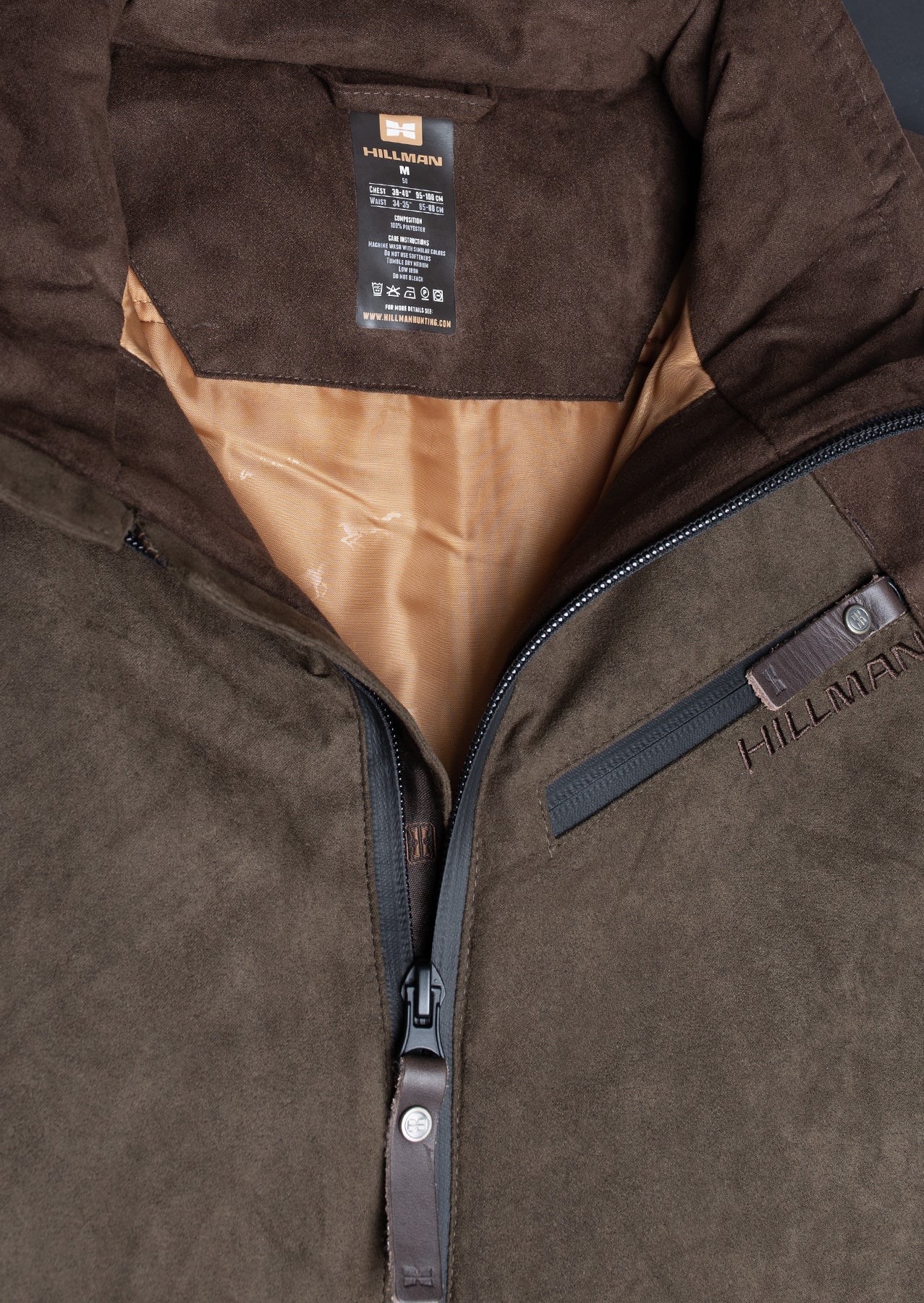 A close-up of the Hillman® Waterproof Insulated Hunting Jacket | Fusion shows a partially open zipper revealing tan lining, a visible care label inside, and detailed views of the zipper pull and chest pocket.