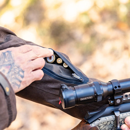 A tattooed hand opens the zippered sleeve pocket of a HILLMAN GEAR Camo Hunting Jacket 6OL Heated - Cold Weather Insulated, revealing rifle cartridges. A scoped, ornate rifle is in the foreground with a blurred background.