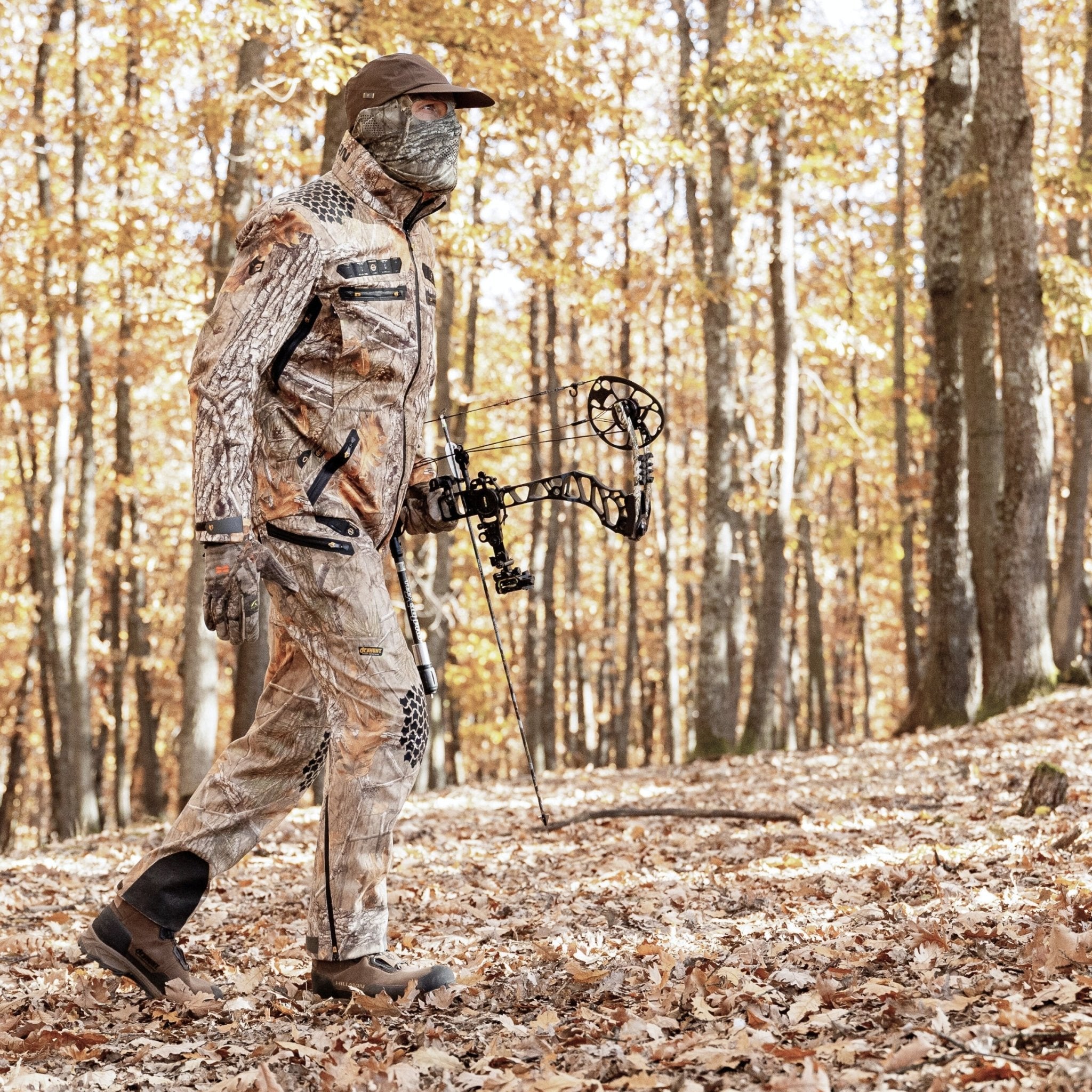 Wearing Hillman® Insulated Hunting Pants Waterproof Cold-Weather ProShell 6OL, a person with a compound bow walks through a forest with autumn foliage and fallen leaves underfoot.