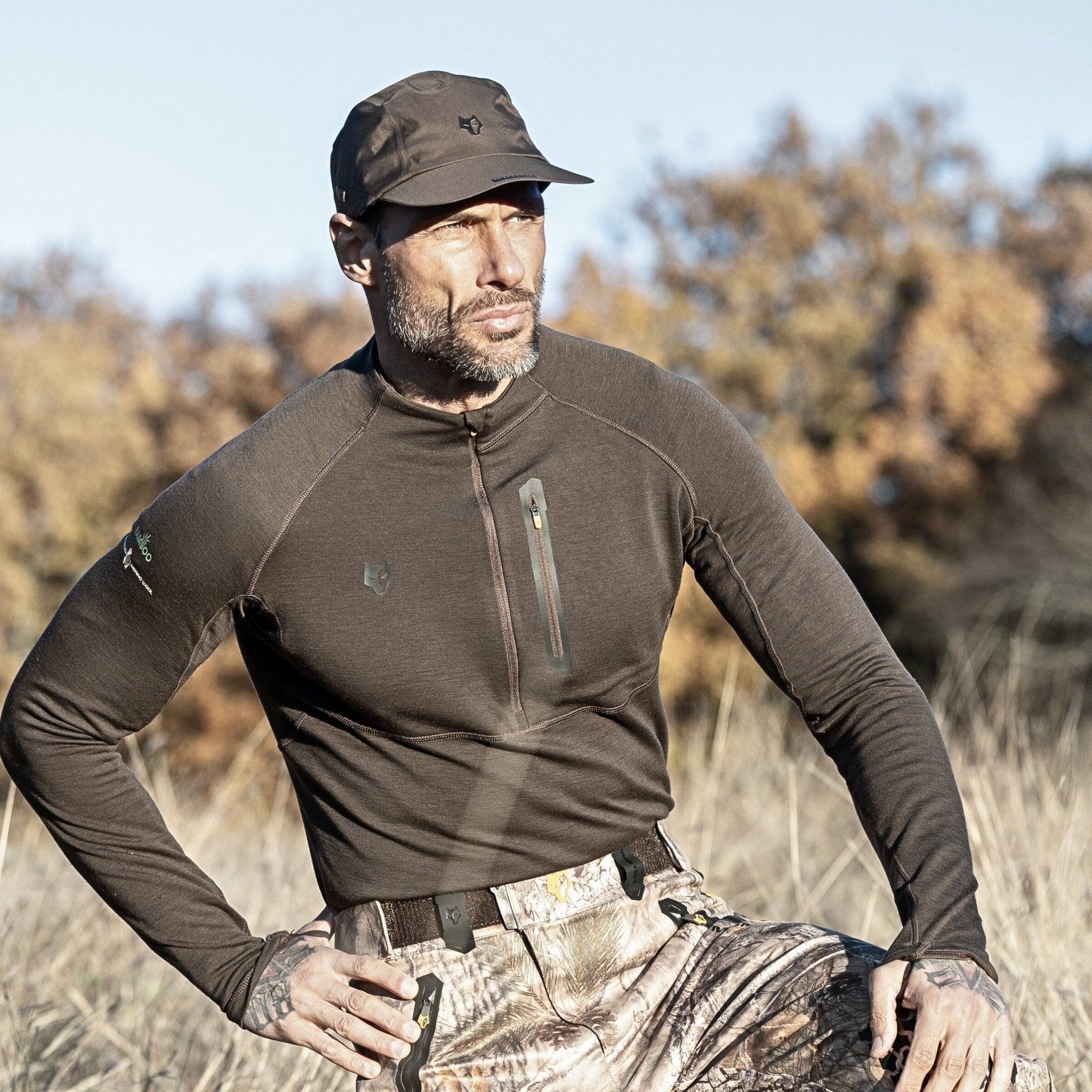 A man in camouflage pants and a Hillman®GWR 2BL Merino Wool Base Layer-Hunting Long Sleeve Thermal T-shirt, with a zippered chest pocket, stands outdoors in a field of dry grass and trees. Wearing a cap, he gazes into the distance, enjoying the shirts thermal regulation for comfort.