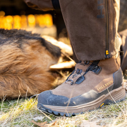 Close-up of a HILLMAN DRYHUNT 2.0 Waterproof Hunting Boot on grass and straw, with a fur coat and person in dark clothing casting shadows nearby. These boots promise comfort for performance hunting adventures.