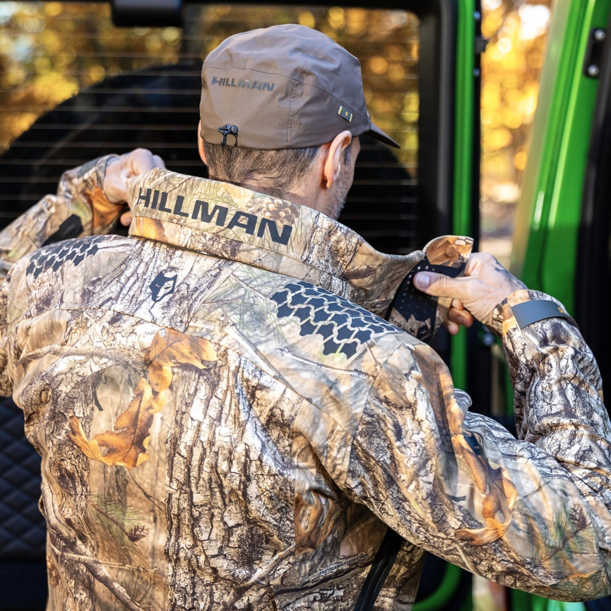 A man in a HILLMAN GEAR Camo Hunting Jacket 6OL Heated - Cold Weather Insulated and cap adjusts his collar next to a green vehicle with an open door, surrounded by autumn scenery ready for outdoor adventure.