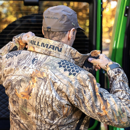 A man in a HILLMAN GEAR Camo Hunting Jacket 6OL Heated - Cold Weather Insulated and cap adjusts his collar next to a green vehicle with an open door, surrounded by autumn scenery ready for outdoor adventure.
