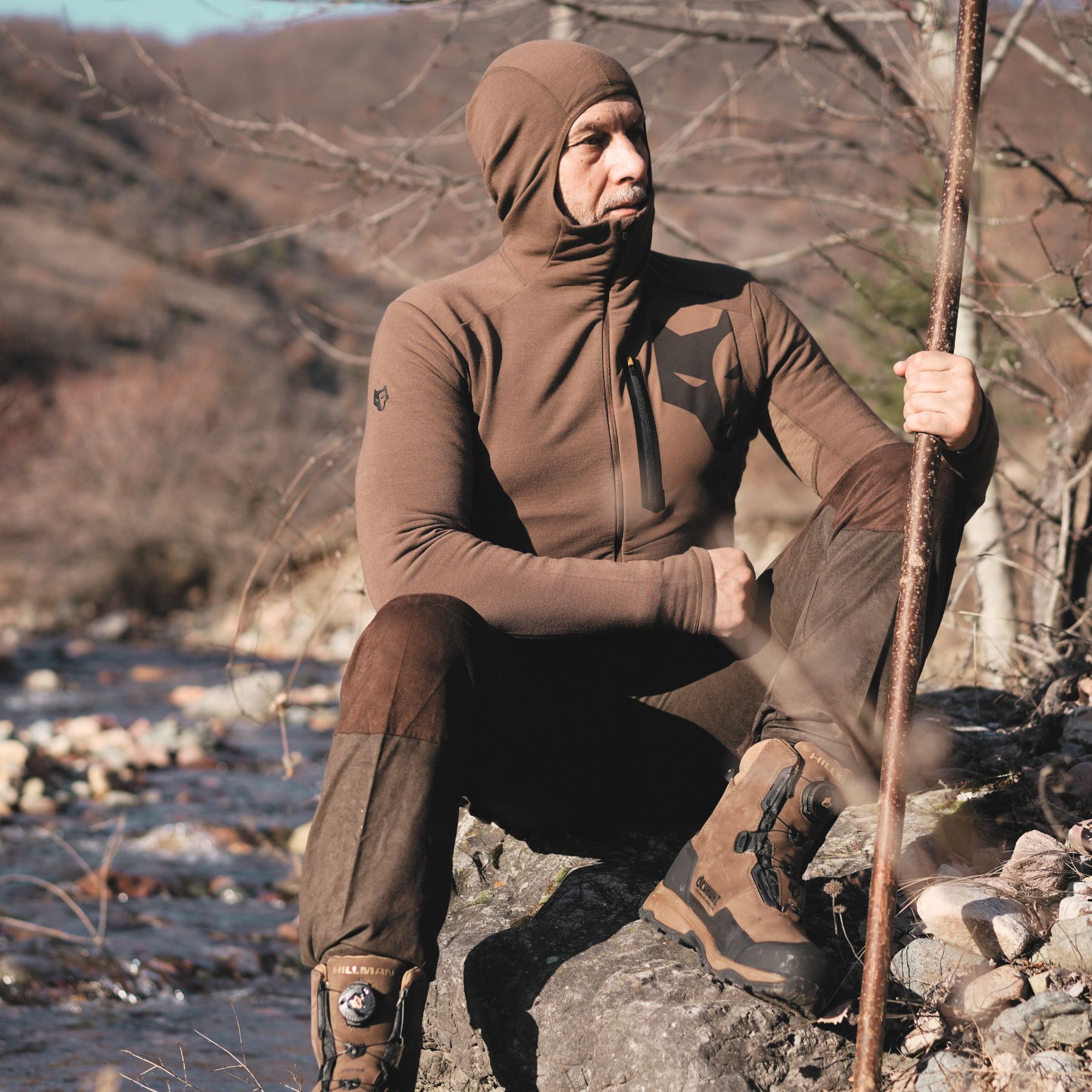 A person sits on a rock by a stream with a wooden stick, wearing the Hillman®GWR 3ML Hunting Hoodie – Warmest Merino Wool Mid Layer for thermal regulation and sturdy hiking boots. Leafless trees and rolling hills form the peaceful backdrop.