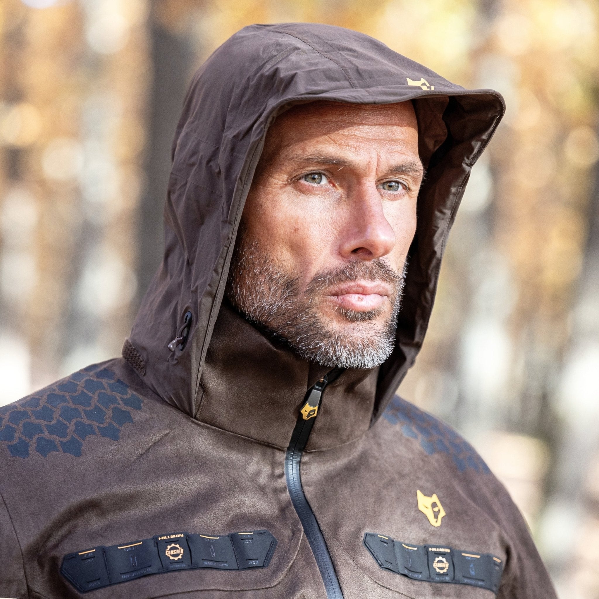 A man with a beard wears a HILLMAN GEAR 5WL Waterproof Camo Hunting Rain Jacket, showcasing its ultra-lightweight design and geometric-patterned shoulders. The jacket features zippers and a logo on the chest against out-of-focus autumn trees with golden leaves.