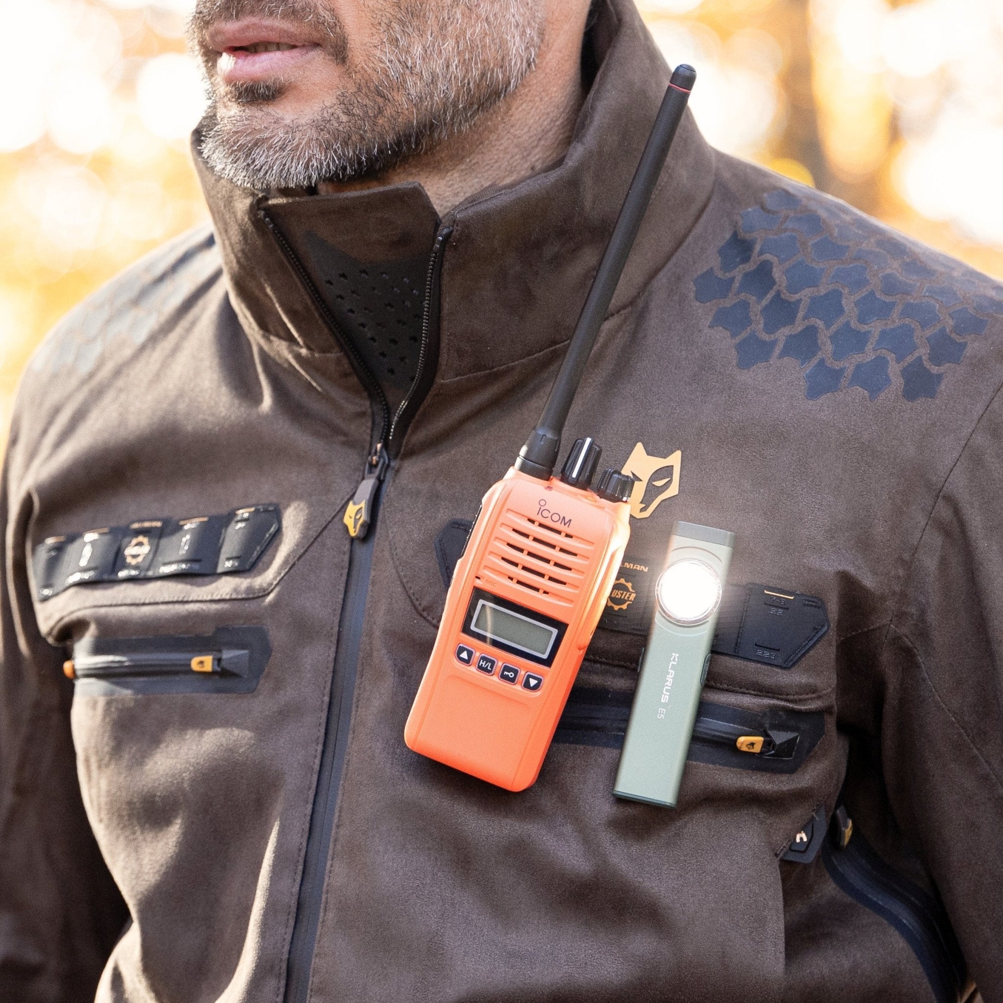A person in a HILLMAN GEAR Camo Hunting Jacket 6OL Heated - Cold Weather Insulated with hexagonal camo, multiple zippers, and utility patches has an orange two-way radio and a green flashlight clipped to the chest.