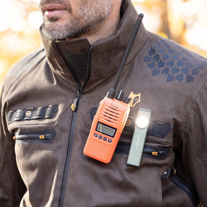 A person in a HILLMAN GEAR Camo Hunting Jacket 6OL Heated - Cold Weather Insulated with hexagonal camo, multiple zippers, and utility patches has an orange two-way radio and a green flashlight clipped to the chest.