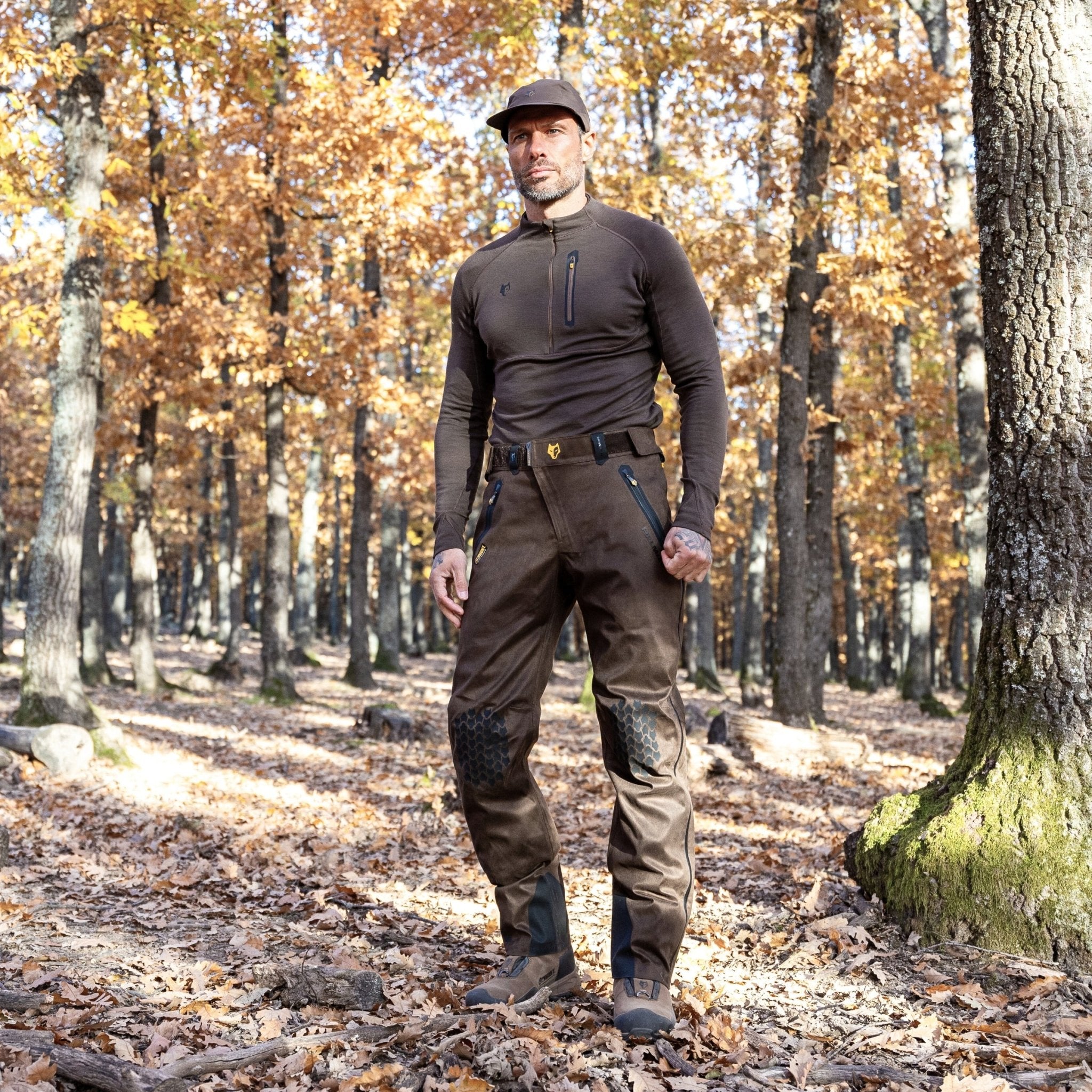 A man stands in a forest with autumn foliage, wearing a brown hat and long-sleeved shirt. His 5WL Waterproof Camo Hunting Pants by HILLMAN GEAR blend seamlessly with the surroundings. Sunlight filters through the trees, casting dappled light on the leaf-covered ground.