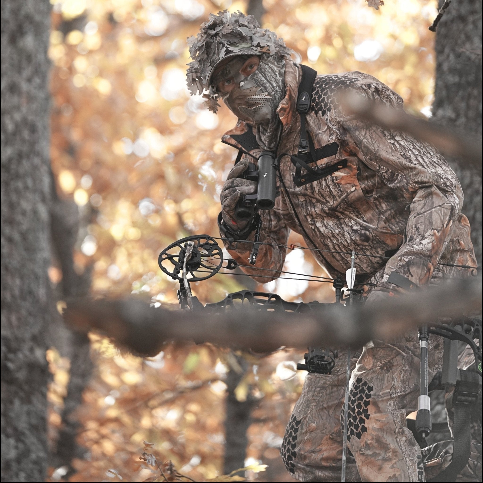 A person wearing the HILLMAN GEAR Camo Hunting Jacket 6OL Heated - Cold Weather Insulated, along with a face covering and hat, stands in an autumn forest holding binoculars and a compound bow.