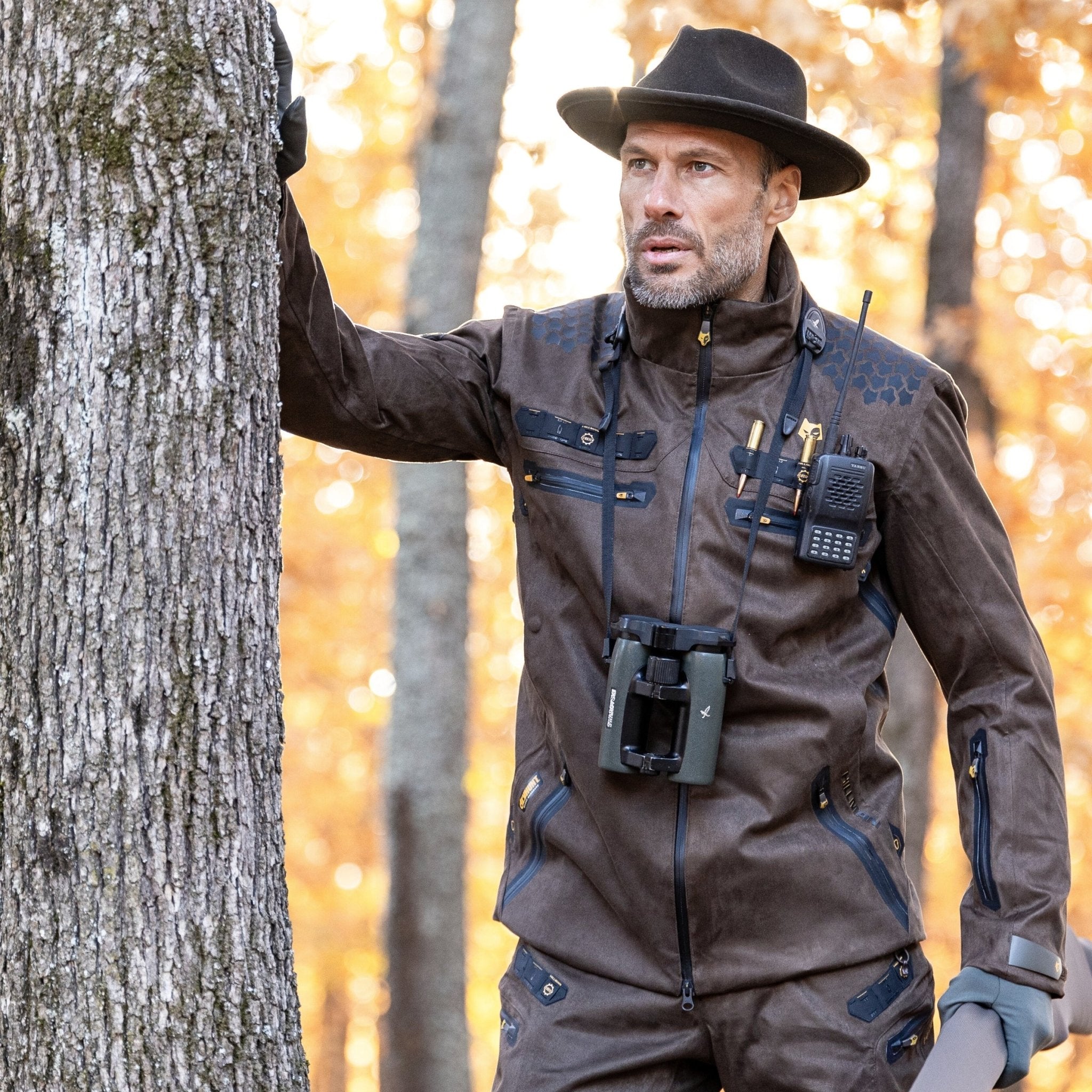 A man in outdoor attire stands in a wooded area, blending into the autumn landscape. He wears a brown 6OL Cold Weather Hunting Jacket by HILLMAN GEAR with multiple pockets, a black hat, and carries binoculars and a two-way radio amid the golden-leaved trees.